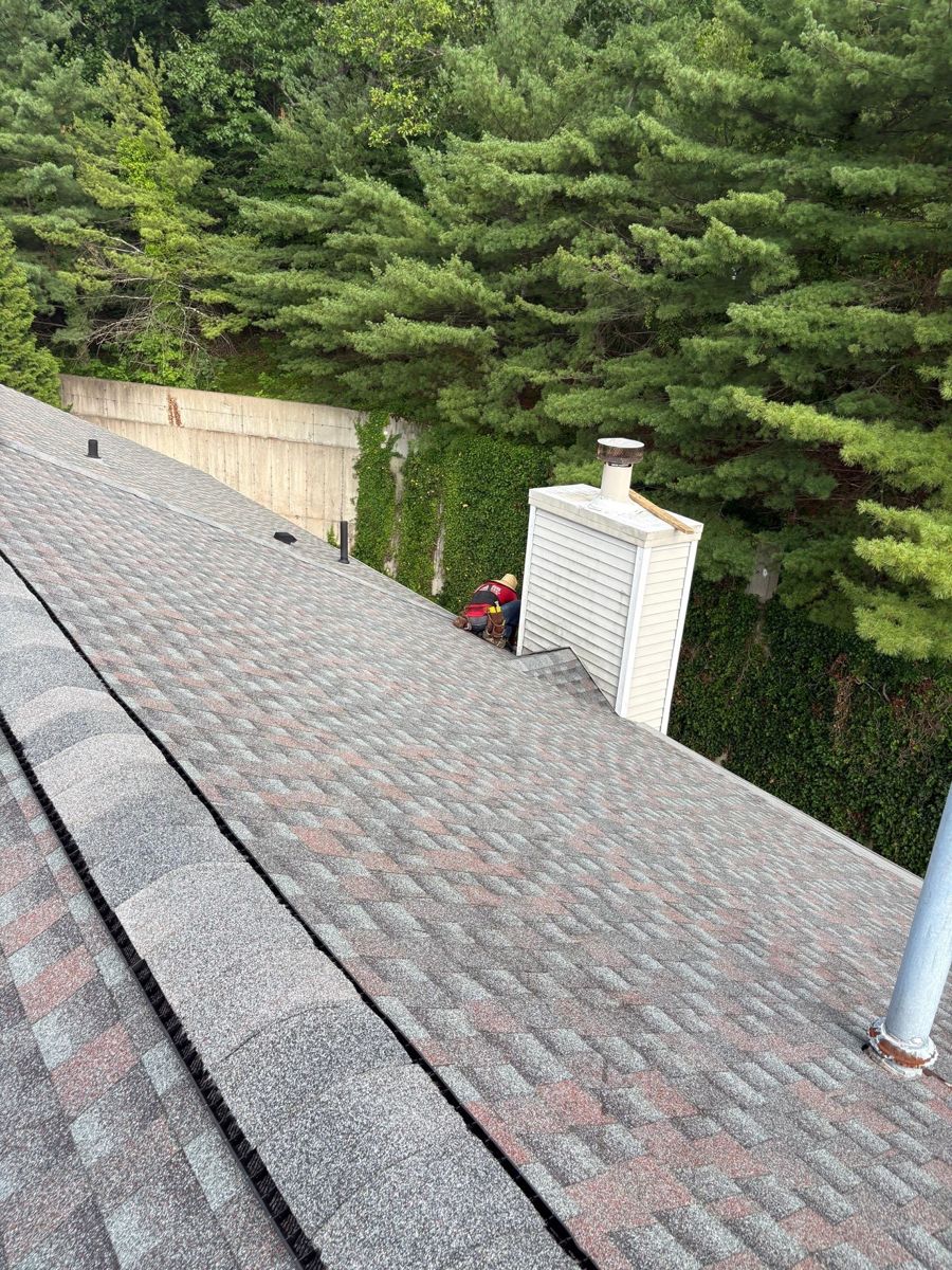 Finished GAF Timberline HDZ roof with ridge vent beside a vinyl chimney chase under tall pines